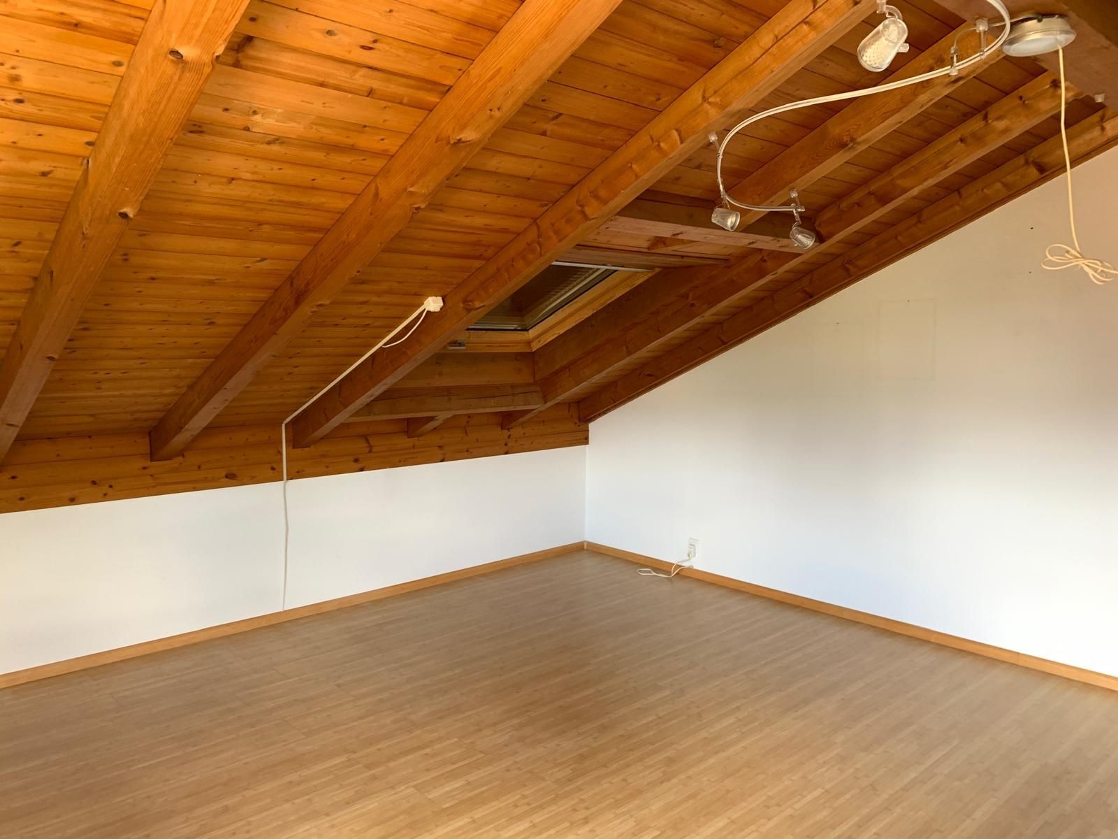 Empty attic room with wooden ceiling beams, white walls, and a light wood floor.